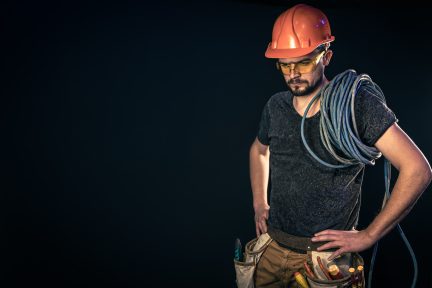 Male electrician in a protective helmet with an electric cable on a black background, copy space.